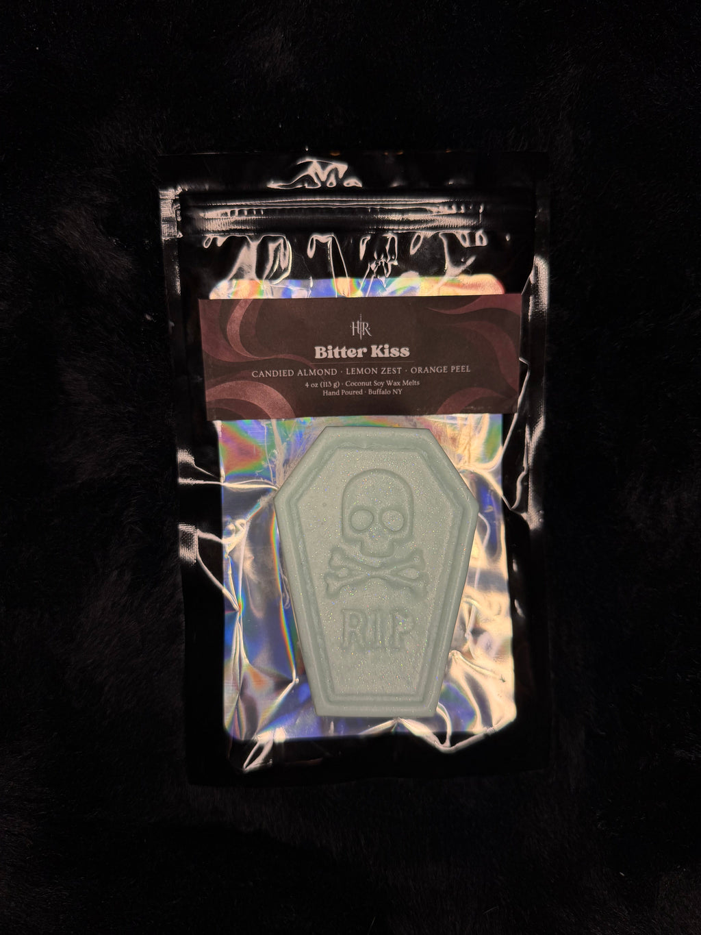 A packaged decorative item on a black furry background. The product appears to be a coffin-shaped pale green object with a skull and crossbones design, sealed inside a shiny holographic pouch with a dark brown label card.