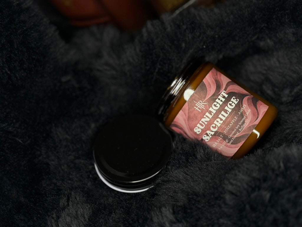 A candle jar labeled 'SUNLIGHT SACRILEGE' with a pink label and amber-brown wax, placed on a dark fuzzy surface next to a black lid.